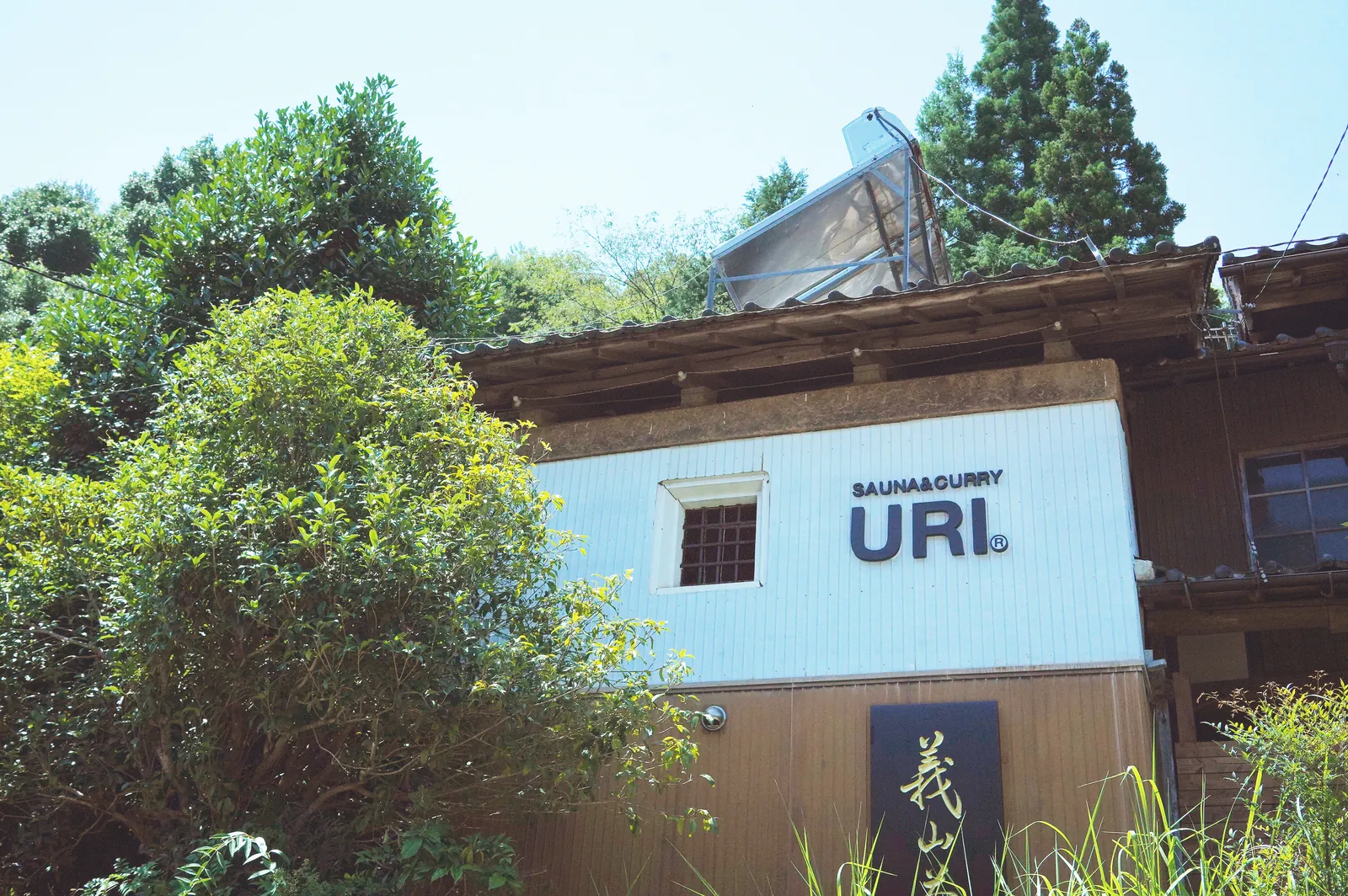 緑の木々の向こうに見える、白壁に「SAUNA&CURRY URI」と書かれた栄の間の看板